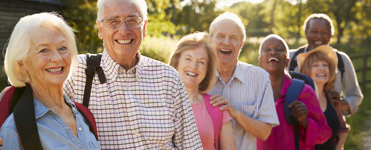 A group of older adults, perhaps ideal for developing buyer personas, stand outdoors in a line, smiling at the camera on a sunny day. Some have backpacks. Trees and sunlight brighten the background.