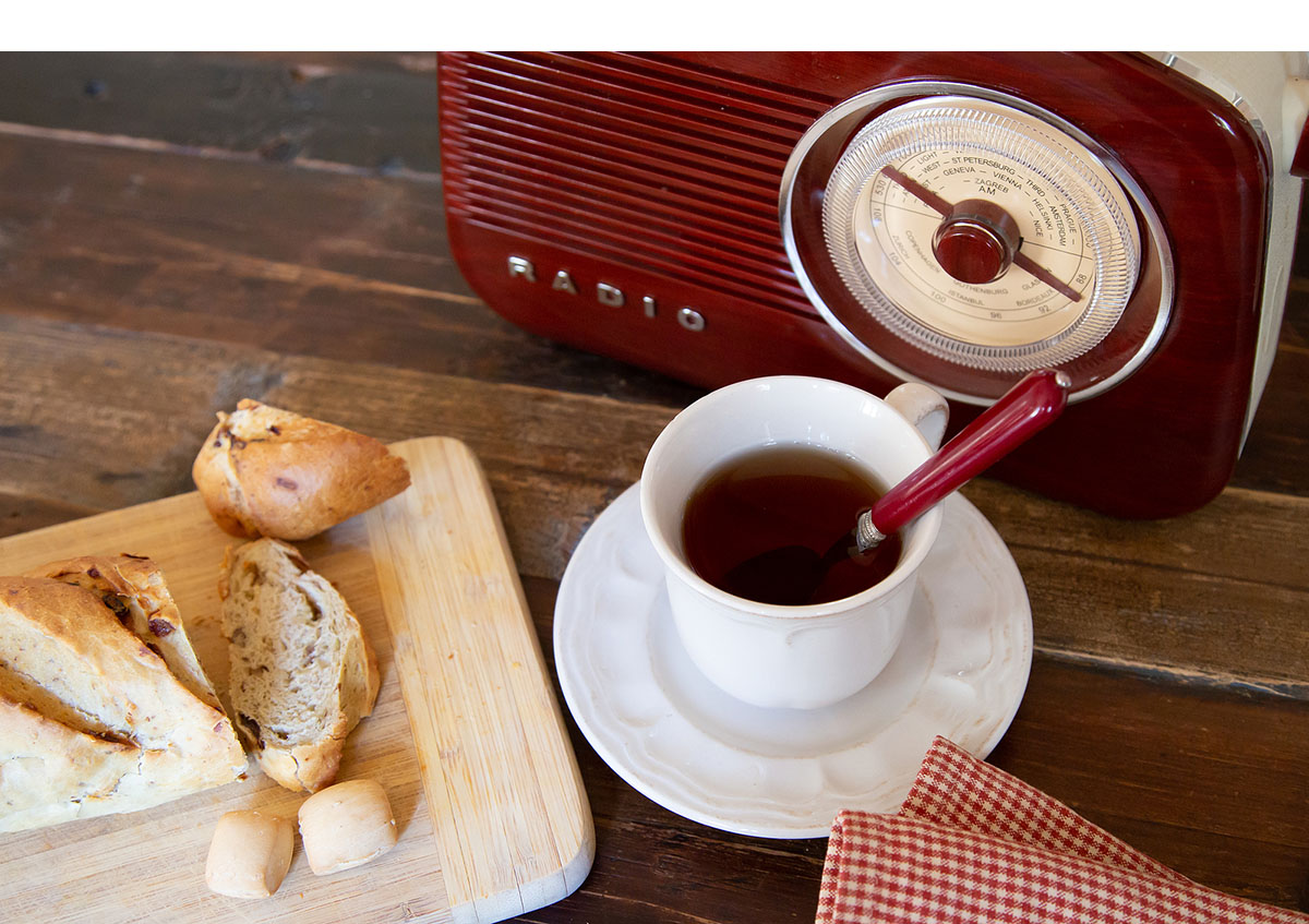 A cup of tea with a spoon on a saucer sits next to sliced bread on a cutting board and a vintage radio, creating an inviting scene perfect for senior living advertising on this cozy wooden table.