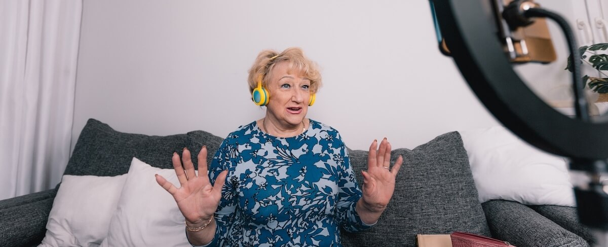 Older woman in a blue floral shirt and yellow headphones sits on a sofa, gesturing with raised hands as she speaks to her smartphone on a ring light—embracing senior living and connecting through social media.