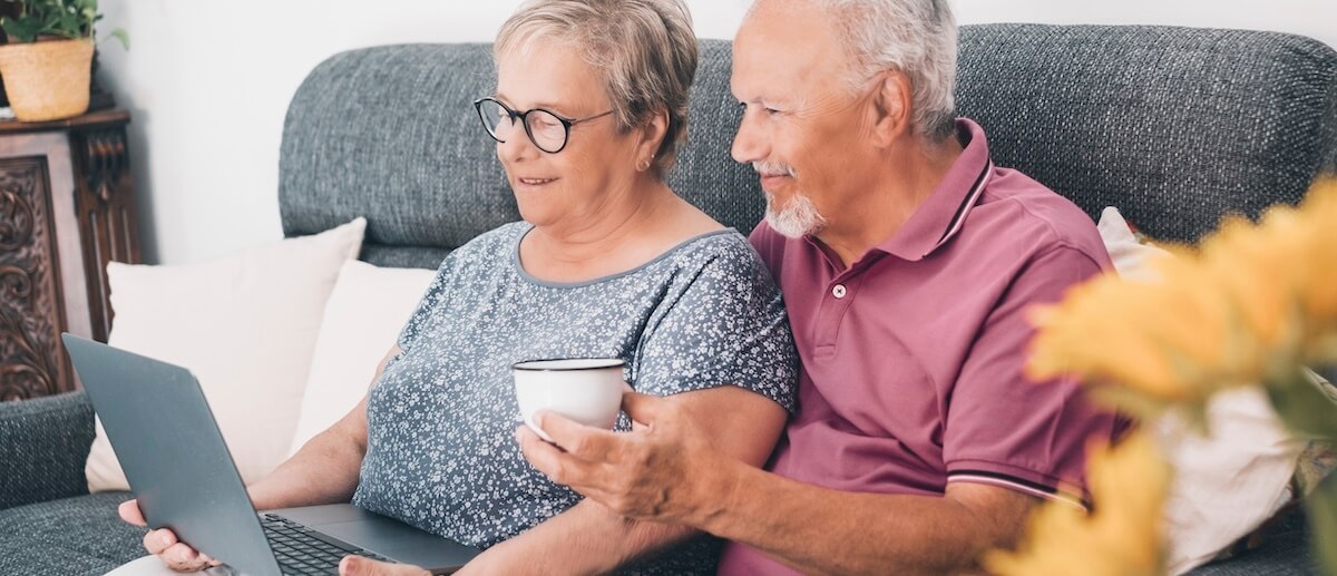 Two older adults sit together on a couch, looking at a laptop screen. One holds a mug, and both appear engaged and content as they browse a senior living website design.