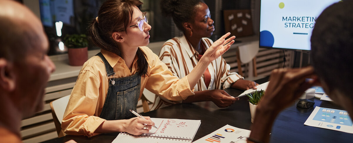 Three people sit at a table with documents and a laptop, discussing a senior living marketing plan in a meeting room. One person gestures while taking notes. A screen displays “Marketing Strategy.”.