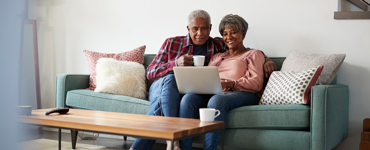 An older couple sits on a sofa, looking at a laptop together—perhaps searching how to market senior living communities. The man holds a mug, and both appear relaxed in their cozy living room setting.