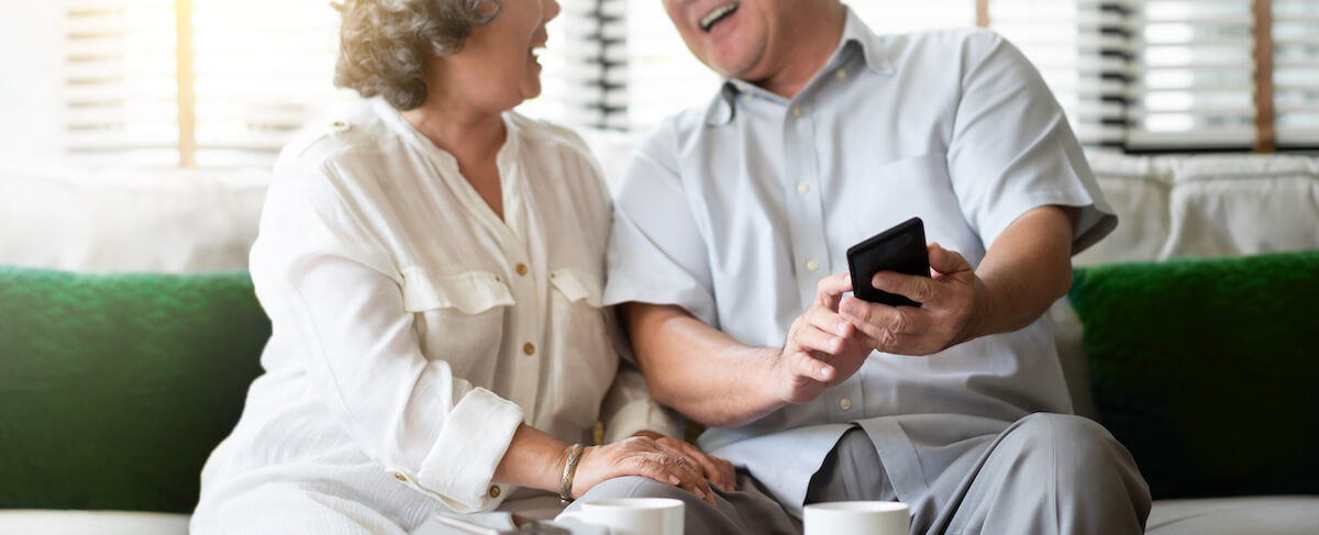 An older couple sits on a sofa, smiling and looking at a smartphone together, possibly exploring advertising on social media, with coffee cups on the table in front of them.