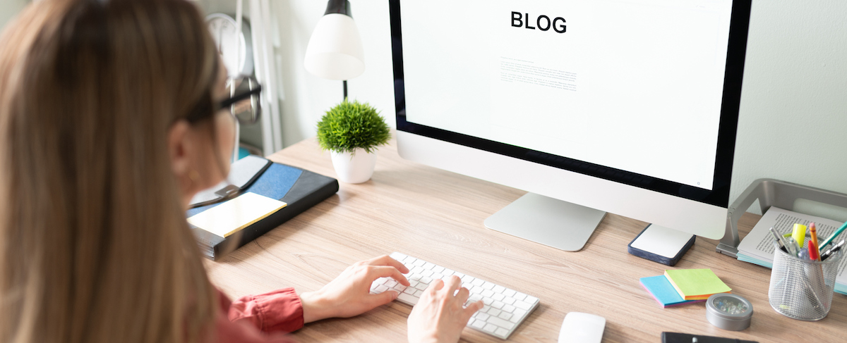 A person sits at a desk, typing on a keyboard in front of a large computer monitor displaying the word "BLOG," brainstorming digital marketing strategies for senior living amid office supplies in a tidy workspace.