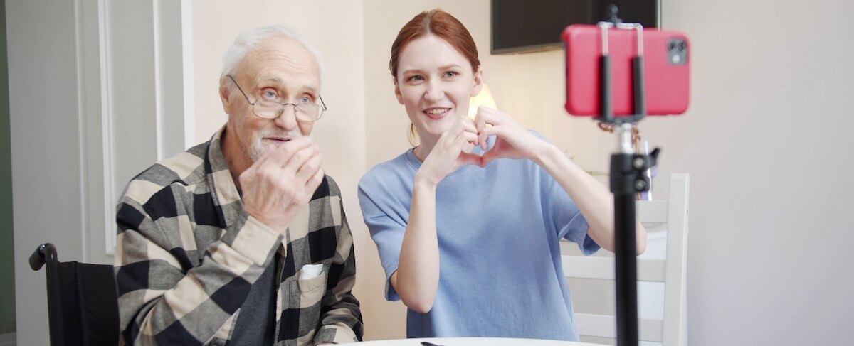 An elderly man and a young woman sit at a table, making hand gestures to a smartphone on a tripod, likely recording or video chatting for video marketing purposes.