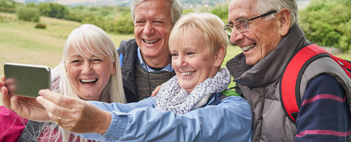 Four older adults stand outdoors taking a selfie together, all smiling and enjoying the moment—perfect inspiration for a senior living website design set among trees and grass in the background.