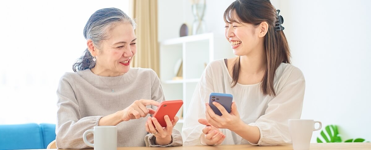 Two women sit at a table with mugs, holding smartphones and smiling at each other in a bright, indoor setting—capturing the warmth and connection that senior living SEO aims to highlight.