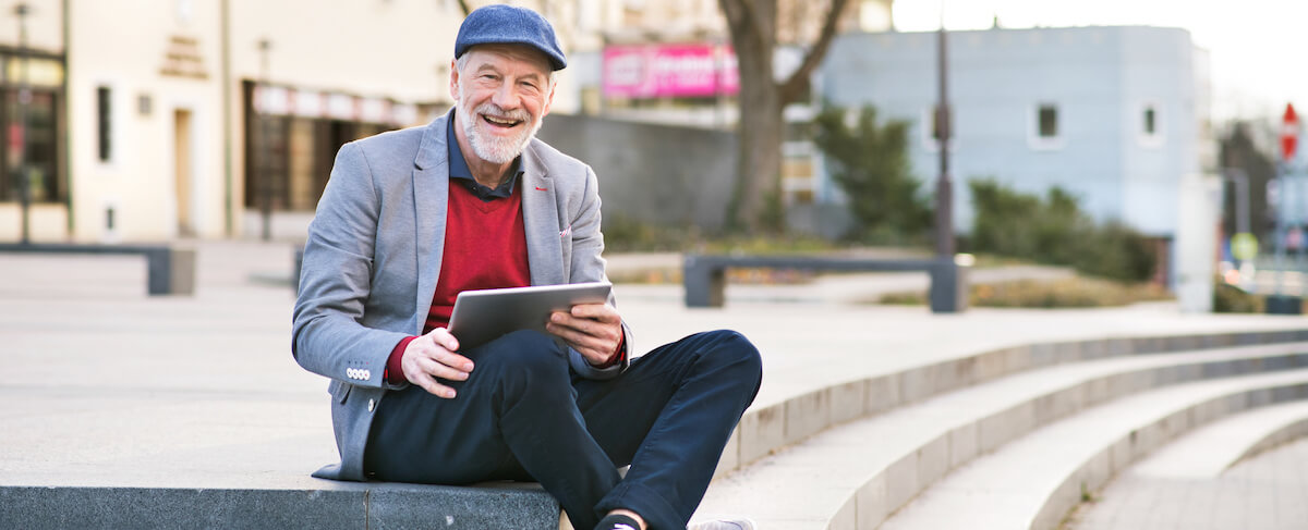 Older man with a gray beard and cap sits on outdoor steps, smiling as he holds a tablet—perhaps exploring the best content for email marketing—in a gray blazer, red sweater, and dark pants.