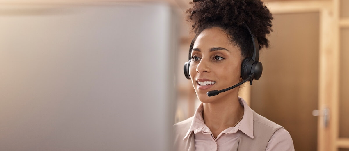 Woman wearing a headset sits in front of a computer monitor, appearing engaged in a Senior Living Marketing conversation or customer service task in an office setting.