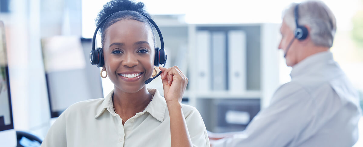 Two customer service representatives wearing headsets work at desks in an office, assisting with senior living leads. The woman in the foreground is smiling at the camera.