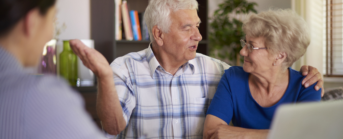 An older man and woman sit together indoors, with the man speaking animatedly and gesturing, while another person listens from across a table—an engaging scene often seen during senior living sales strategies discussions.