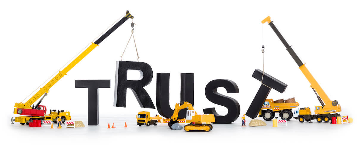 Construction vehicles assembling large black letters spelling "TRUST" on a white background, visually illustrating how to build trust with prospects as cranes lift each letter into place.