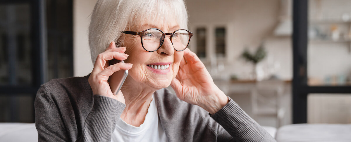 Elderly woman with short white hair and glasses smiling while talking on a smartphone, sitting indoors in a well-lit room—perhaps practicing senior living sales training.
