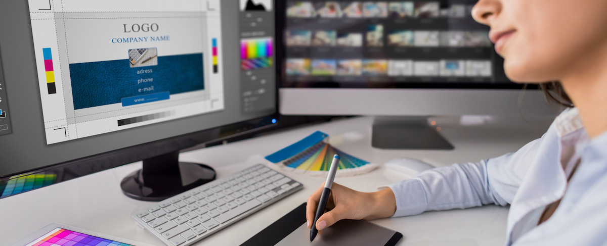 Person using a graphics tablet to design a company logo and business card on a computer, with color swatches, editing software, and inspiration from the best senior living ads visible on the desk.