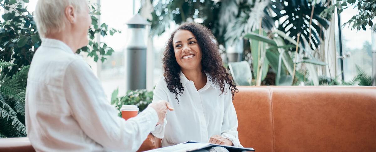Two people sitting on a couch in a bright, plant-filled space, smiling and shaking hands over papers and coffee—demonstrating how to engage customers through warm, personal connections.