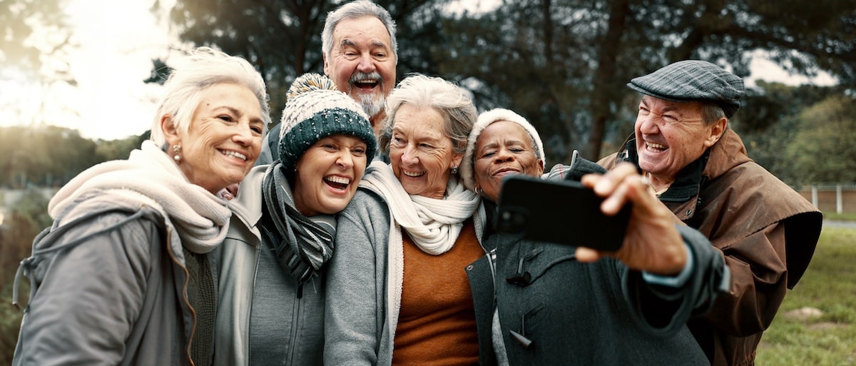 Six older adults wearing winter clothing stand outdoors, smiling and posing closely together as one person takes a selfie with a smartphone—capturing authentic moments perfect for senior living social media marketing.