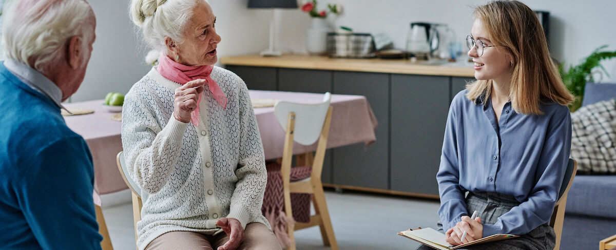 Three people sit indoors having a conversation; an elderly woman gestures while speaking, an elderly man listens, and a younger woman with a notepad—there for memory care lead generation—smiles and observes.