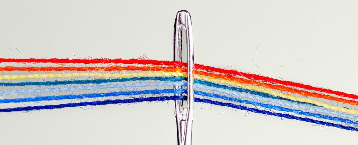 Close-up of multiple colorful threads, including red, yellow, blue, and orange—perfect threads for marketing—threaded through the eye of a sewing needle against a plain background.