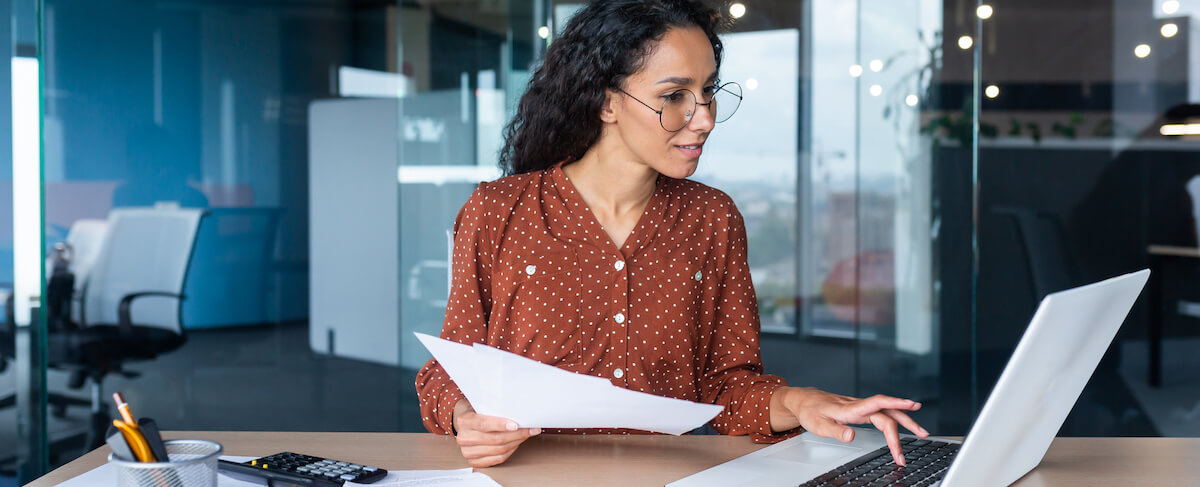 Woman wearing glasses sits at a desk in an office, holding papers in one hand and typing marketing analytics data into a laptop with the other.
