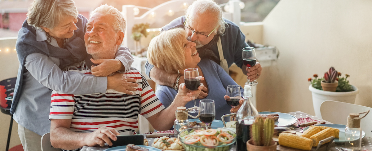 Four older adults sit at a table outdoors, sharing a meal and drinks, with two couples embracing and smiling at each other—an inspiring scene for creative marketing ideas for senior living communities.
