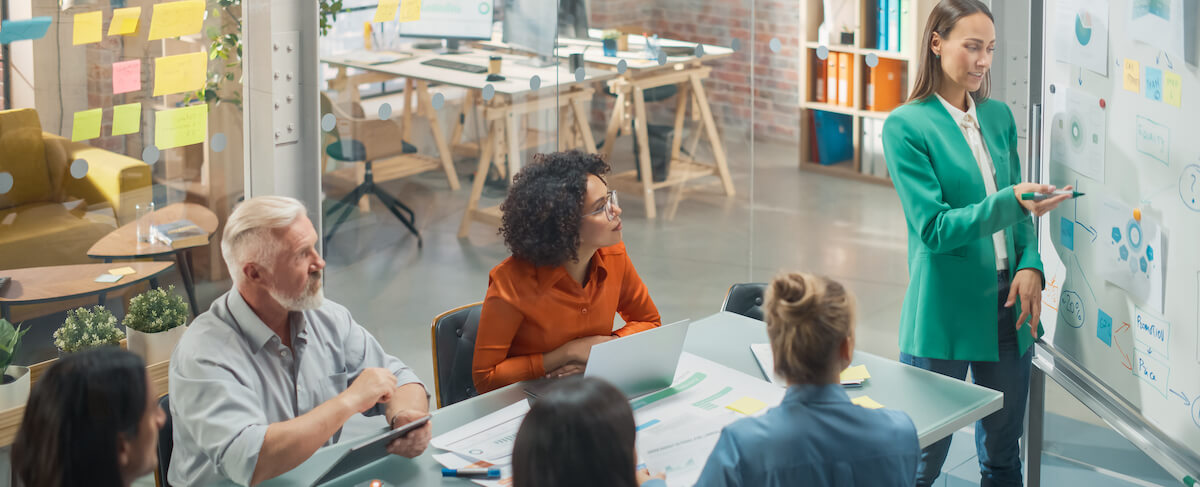 A woman stands at a whiteboard presenting data on senior living marketing budgets to a group of five colleagues seated around a table in a modern office setting.