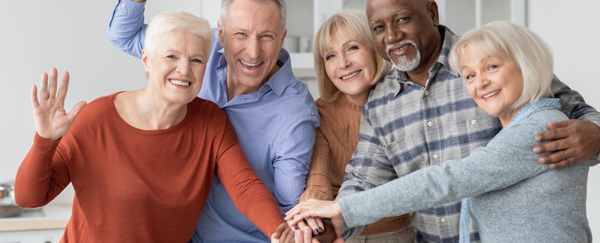 Five older adults stand close together indoors, smiling at the camera with hands stacked in unity—one person waves. A perfect snapshot for any senior living guide celebrating community and connection.