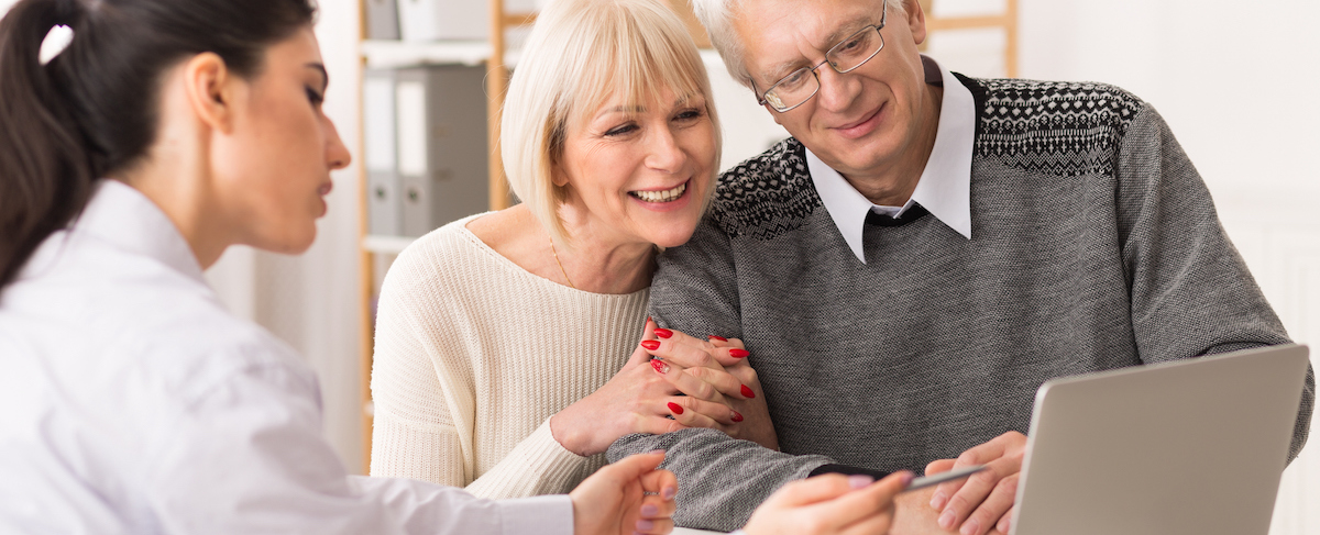 A senior couple sits together, listening to a professional woman explain senior living sales options on a laptop in an office setting.
