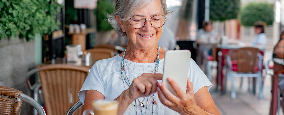 Older woman with short gray hair and glasses smiles while using her smartphone at an outdoor café, likely browsing senior living PPC options as she enjoys a coffee in front of her.