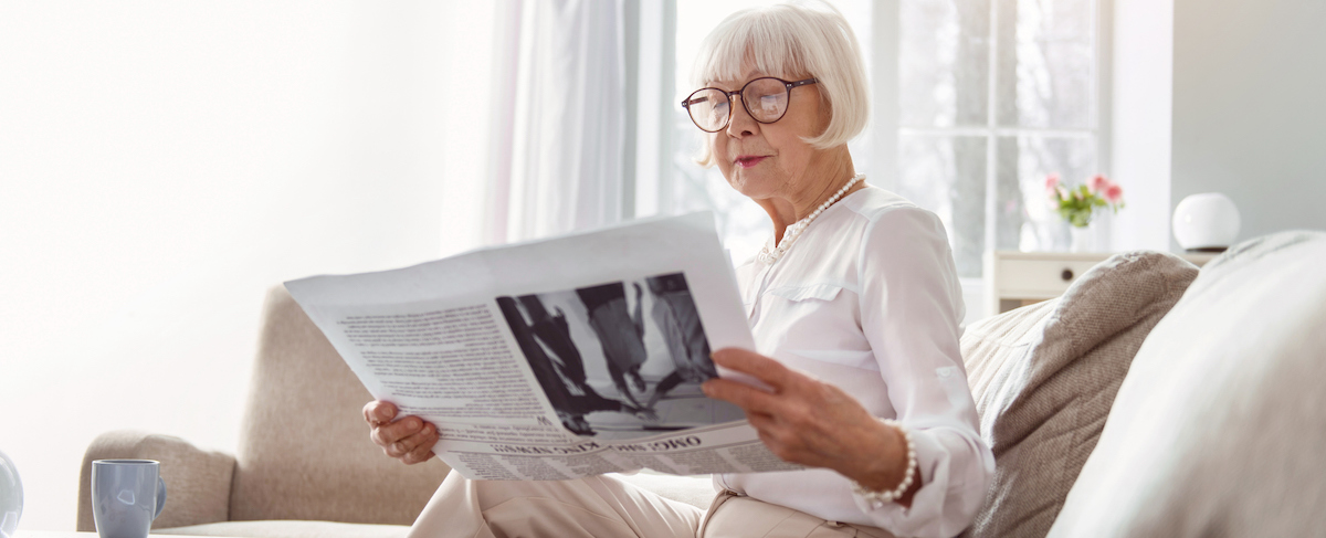 Elderly woman with glasses sits on a sofa, reading a newspaper in a bright living room with a window and flowers—an ideal scene for a senior living marketing strategy.