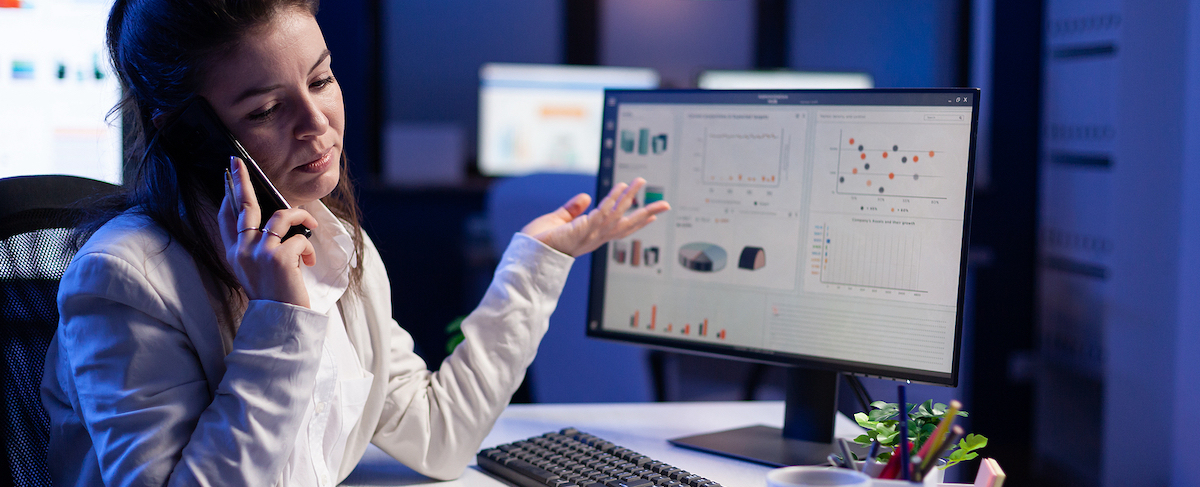 A woman in business attire talks on the phone at her desk, gesturing toward a computer monitor displaying charts and graphs related to senior living marketing software in an office setting.