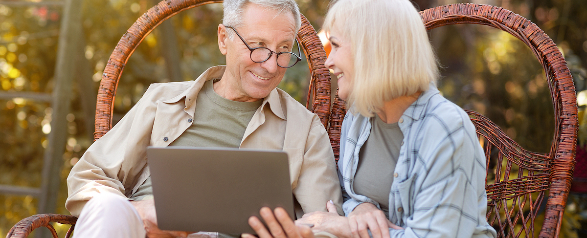 Two older adults sit side by side on wicker chairs outdoors, smiling as they explore effective email marketing strategies together on a laptop computer.