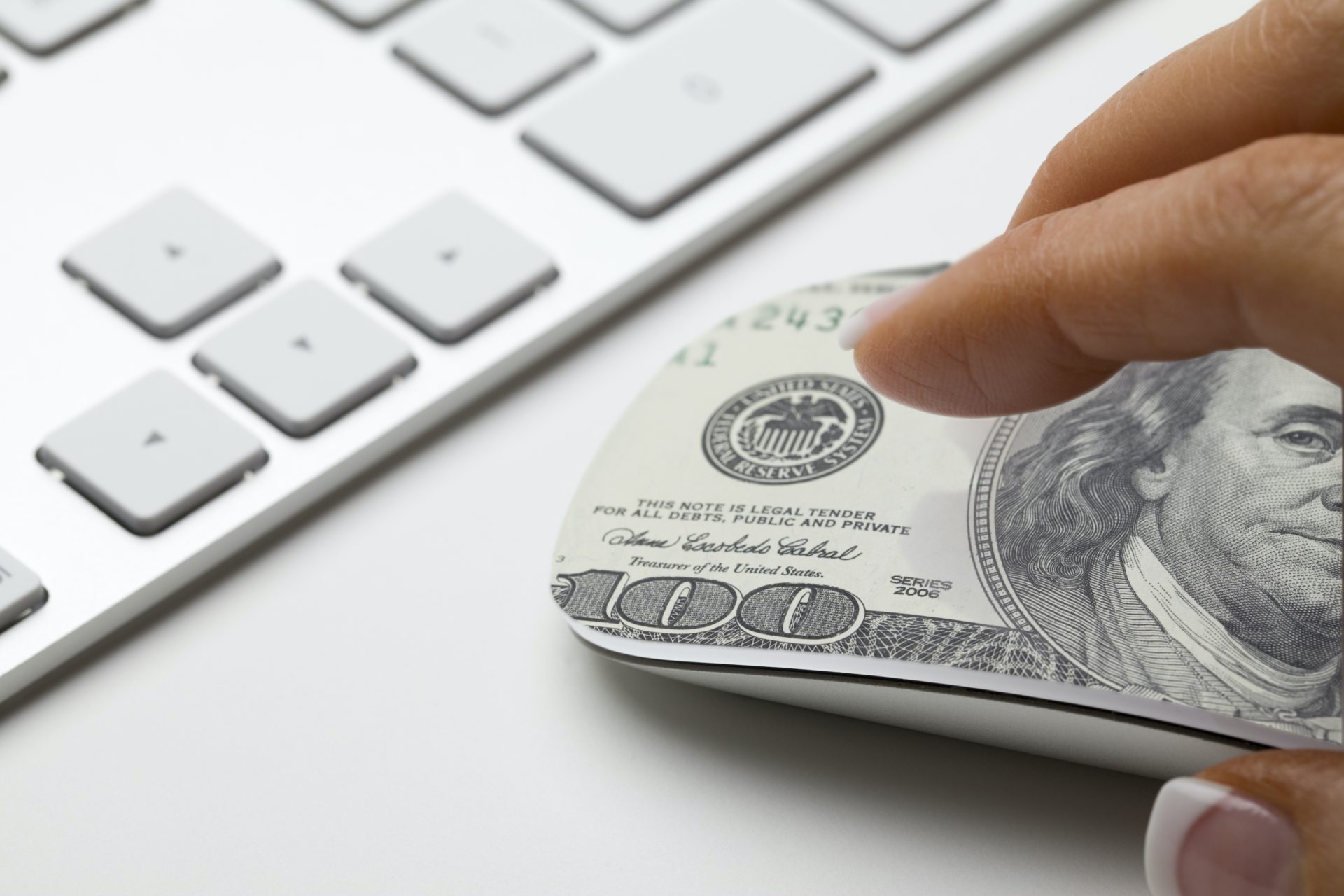 A hand holding a computer mouse wrapped with a US $100 bill, next to a white keyboard on a desk—perfect imagery for senior living advertising focused on the value of smart technology investments.
