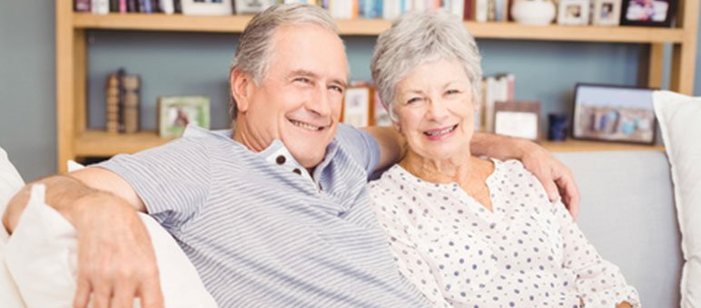 Older man and woman sit close together on a white couch, smiling at the camera—a warm moment that reflects comfort and connection, ideal for senior living sales. Shelves and framed photos add a cozy touch to the background.