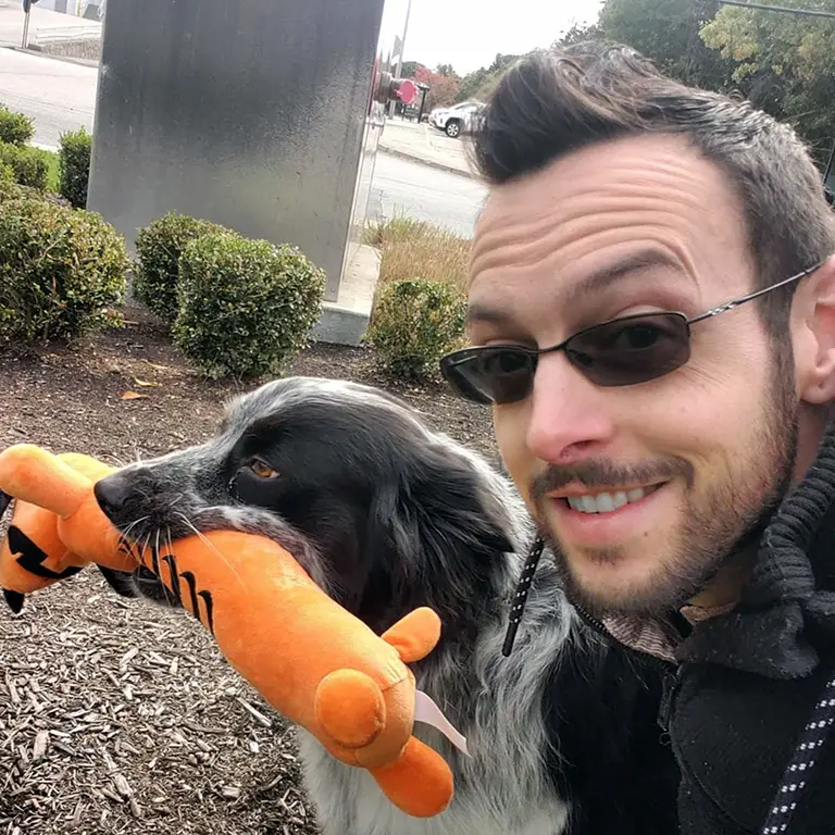 Man wearing sunglasses takes a selfie outdoors with a black and white dog holding an orange plush toy in its mouth.