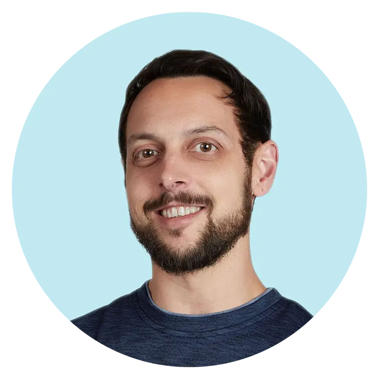 Smiling man with short dark hair and a beard, wearing a blue shirt, posed against a light blue background.
