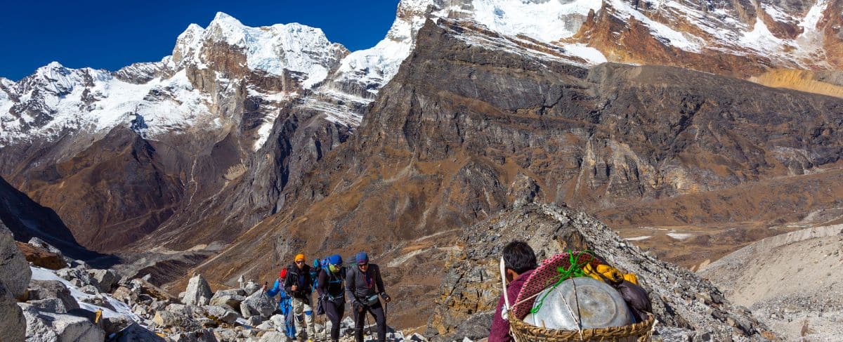 A group of trekkers and a porter with a basket hike along a rocky mountain trail, while snow-covered peaks rise in the background—an inspiring scene reminiscent of adventures featured in senior living marketing.