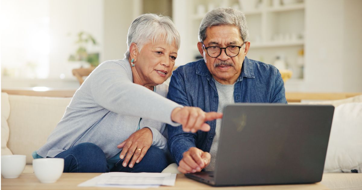 An older woman points at a laptop screen while sitting next to an older man; both are focused on learning Senior Living Sales tips together in a cozy home setting.
