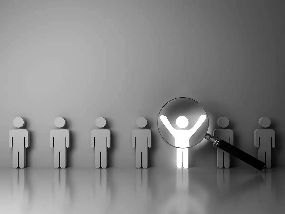 A row of gray human figures with one white figure highlighted by a magnifying glass, symbolizing selection or standing out from the crowd.