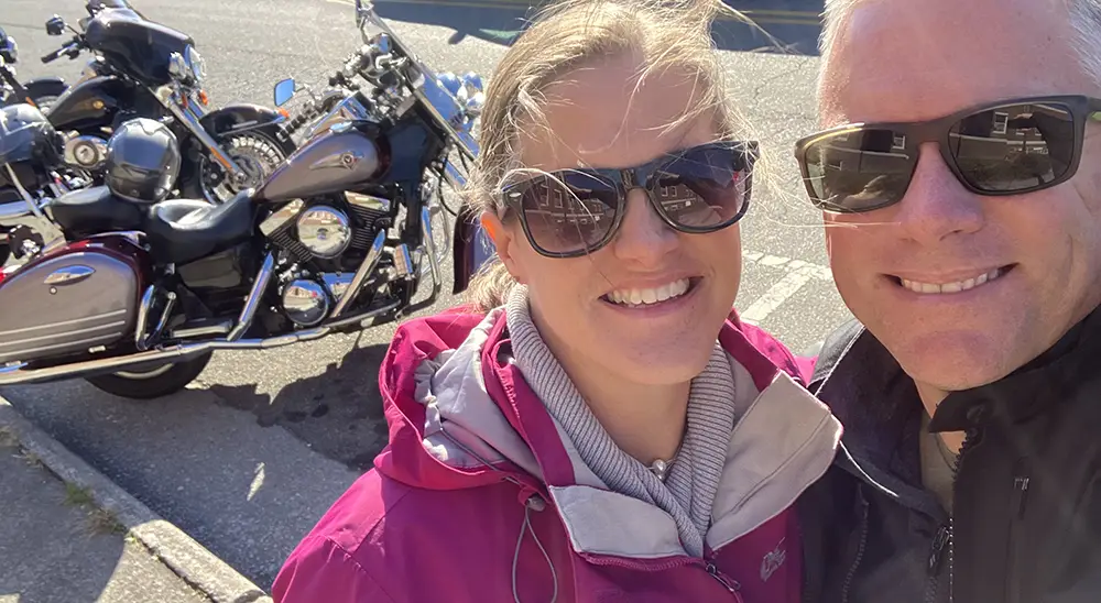 Two people smile for a selfie outdoors with motorcycles parked on the street behind them.