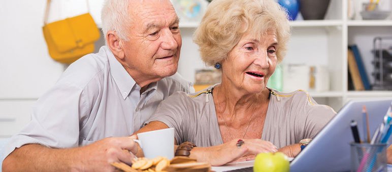 An elderly couple sits at a table, looking at a laptop together. Exploring senior living sales options, the woman touches the keyboard while the man holds a mug. Snacks and an apple are on the table.
