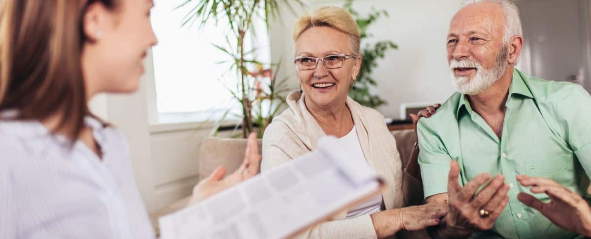 An older couple sits smiling and talking with a woman holding a clipboard, in a bright living room with plants in the background—discussing how SMARTest Marketing uses AI in marketing to reach new customers.