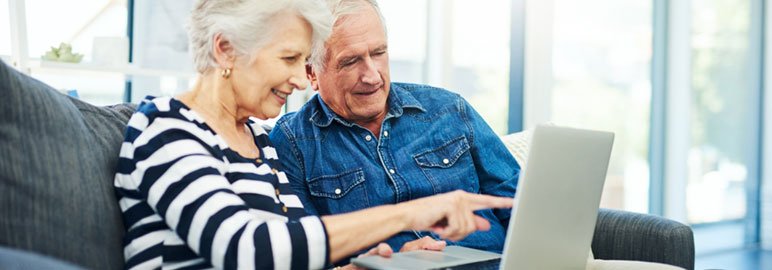 Two older adults sit together on a couch, looking at a laptop screen. One person is pointing at the laptop while both appear focused and engaged, exploring the best senior living websites for helpful resources.