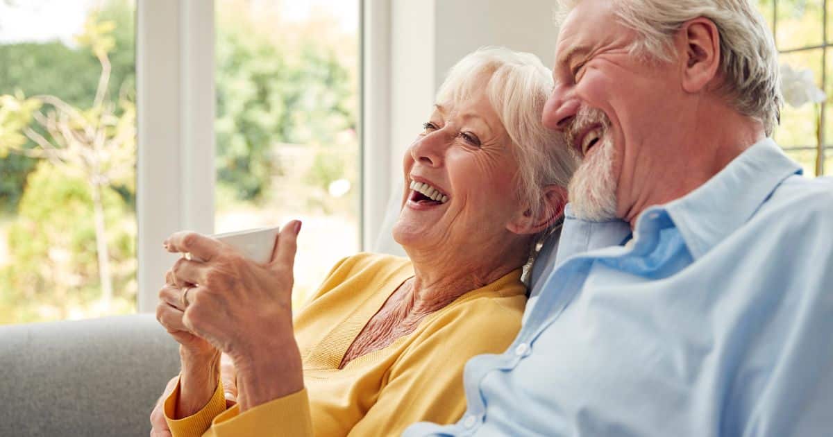 An older woman and man sit close together on a couch, smiling and laughing as they discuss how to manage a waiting list. The woman is holding a white mug while sunlight streams in through large windows behind them.