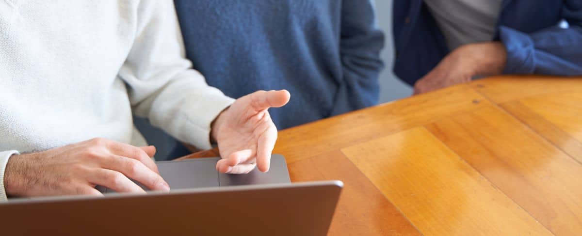 Three people sit at a wooden table, with one person gesturing while using a laptop—perhaps discussing strategies to generate senior living leads. Only their torsos and hands are visible.