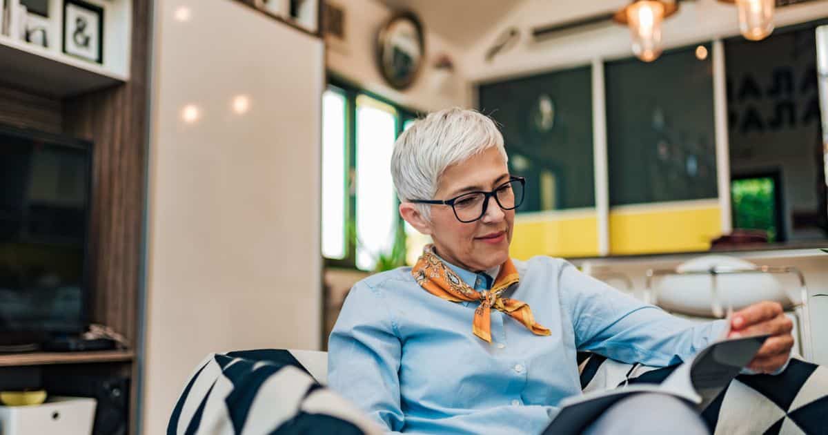 Older woman with short gray hair and glasses sits indoors, reading a magazine—a classic piece of traditional marketing—while holding a drink, wearing a light blue shirt and an orange scarf.