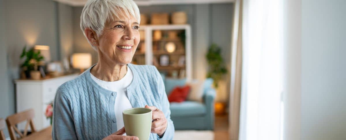 Older woman with short gray hair, wearing a blue sweater, holds a mug and smiles while standing in a bright living room, embodying the comfort smart operators provide to boost occupancy and enhance residents’ happiness.