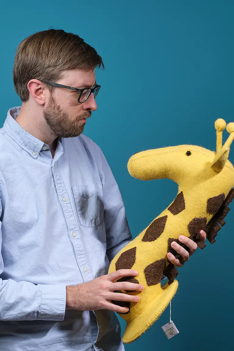 A man wearing glasses and a light blue shirt holds and looks at a large yellow and brown stuffed giraffe toy against a blue background.