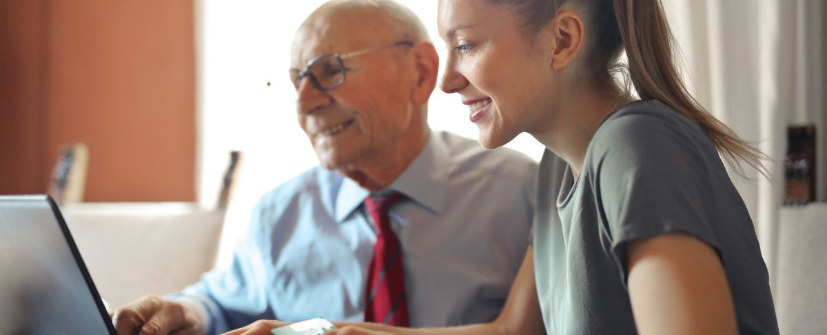 A young woman and an older man sit together at a table, both smiling as they look at a laptop screen, exploring google ads for senior living.
