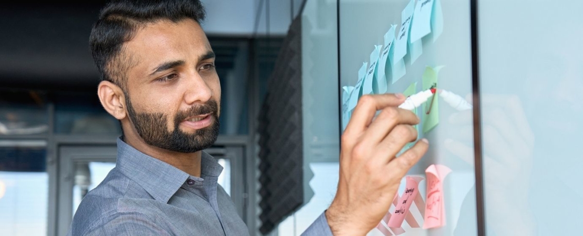Man in a grey shirt writes on a glass board covered with colorful sticky notes in an office, brainstorming ideas on how to increase sales in senior living.