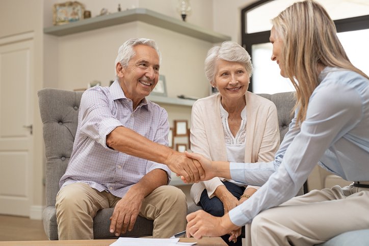 An older couple sits on a couch, smiling and shaking hands with a woman across from them, as they discuss senior living sales strategies in a brightly lit living room.